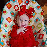 Bailey is registered to the contest to win money with this photo: baby, infant, smiling, red_bow, red_dress, headband, bouncer, baby_seat, patterned_background, geometric_pattern, circles, squares, blue_accent, orange_accent, green_accent, white_background, hand, eyes, indoor, fabric