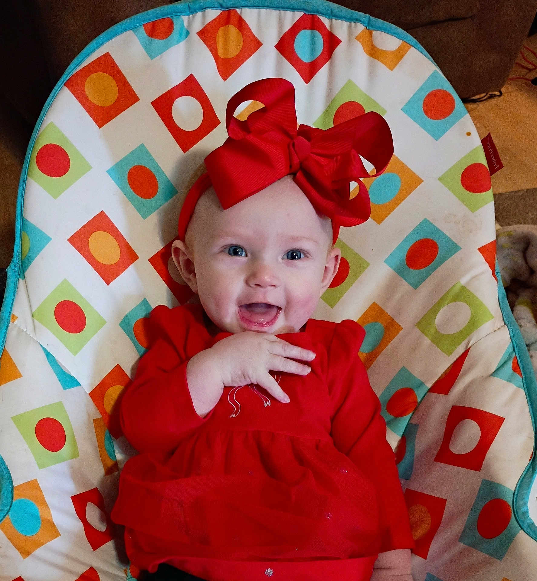Bailey is registered to the contest to win money with this photo: baby, infant, smiling, red_bow, red_dress, headband, bouncer, baby_seat, patterned_background, geometric_pattern, circles, squares, blue_accent, orange_accent, green_accent, white_background, hand, eyes, indoor, fabric