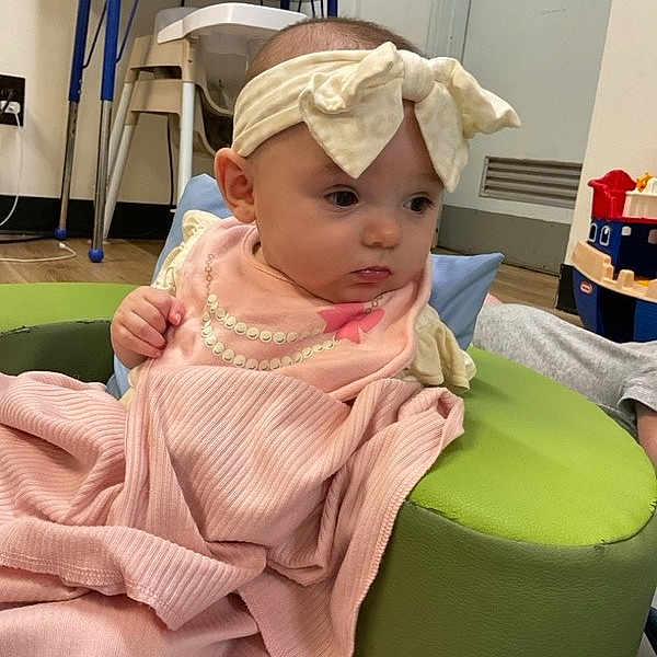 Azeleigh is registered to the contest to win money with this photo: baby, blanket, bow, child, curious, cushion, expression, floor, flooring, furniture, headband, indoor, infant, person, pink_clothing, portrait, room, seated, soft_texture, toy