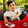 boy, child, driver, face, happy, outdoor, park, pilot_costume, playground, portrait, red_car, sitting, smiling, steering_wheel, sunlight, toddler, toy_car, trees, uniform, vehicle