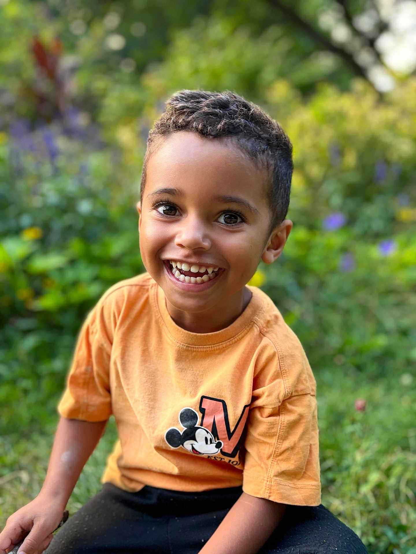 Kaïs a rejoint le concours — aidez-le/la à gagner de superbes lots ! child, boy, smiling, outdoor, garden, greenery, mickey_mouse, tshirt, short_sleeve, happy, cute, portrait, young, casual_clothing, nature, daylight, sitting, curly_hair, playful, bright