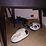 cat, panda_slippers, white_slipper, blue_croc, table, table_leg, chair_leg, floor, wall, electrical_outlet, charger, cable, plush_toy, pet, hiding, shadow, indoor, flash_glare, cozy, shoe