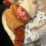 Ange a rejoint le concours — aidez-le/la à gagner de superbes lots ! baby, infant, hat, brown_clothing, blanket, crib, patterned_blanket, hand, face, newborn, cute, soft_texture, indoors, sleeping_area, child, person, resting, closeup, warm_clothing, expression