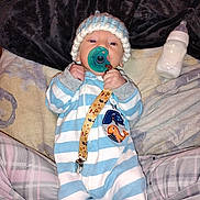 Kane is registered to the contest to win money with this photo: baby, baby_bottle, bed, bedding, blanket, blue, child, cozy, cushions, indoor, infant, knitted_hat, lying_down, onesie, pacifier, portrait, sleepwear, striped_clothing, toy_strap, white