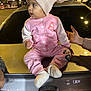 toddler, child, pink_clothing, hat, knit_hat, pom_pom, car, trunk, night, streetlights, hand, adult_hand, parking_lot, license_plate, curious, socks, jacket, pants, outdoor, portrait