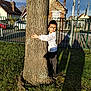 child, boy, tree, glasses, hugging, grass, shadow, house, car, fence, outdoor, sunlight, casual_clothing, smiling, side_view, daytime, nature, yard, residential_area, person