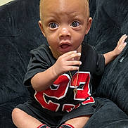 Nico Valmir is registered to the contest to win money with this photo: baby, child, sitting, black_shirt, red_number, necklace, socks, chair, indoor, cute, wide_eyes, expression, plush, comfortable, young, person, portrait, candid, curious, hand