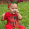 baby, child, grass, outdoor, red_clothing, reindeer_antlers, holiday_theme, smiling, cute, festive, sitting, greenery, tongue_out, playful, infant, nature, headband, joyful, happy, seasonal
