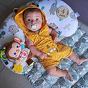 Joseph a rejoint le concours — aidez-le/la à gagner de superbes lots ! baby, infant, pacifier, yellow_clothing, hooded_romper, plush_toy, monkey_toy, floor_mat, leaf_pattern, wooden_plaque, age_marker, indoor, tile_floor, cute, child, young_child, lying_down, wide_eyes, baby_girl, baby_boy