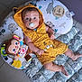 baby, infant, pacifier, yellow_clothing, hooded_romper, plush_toy, monkey_toy, floor_mat, leaf_pattern, wooden_plaque, age_marker, indoor, tile_floor, cute, child, young_child, lying_down, wide_eyes, baby_girl, baby_boy