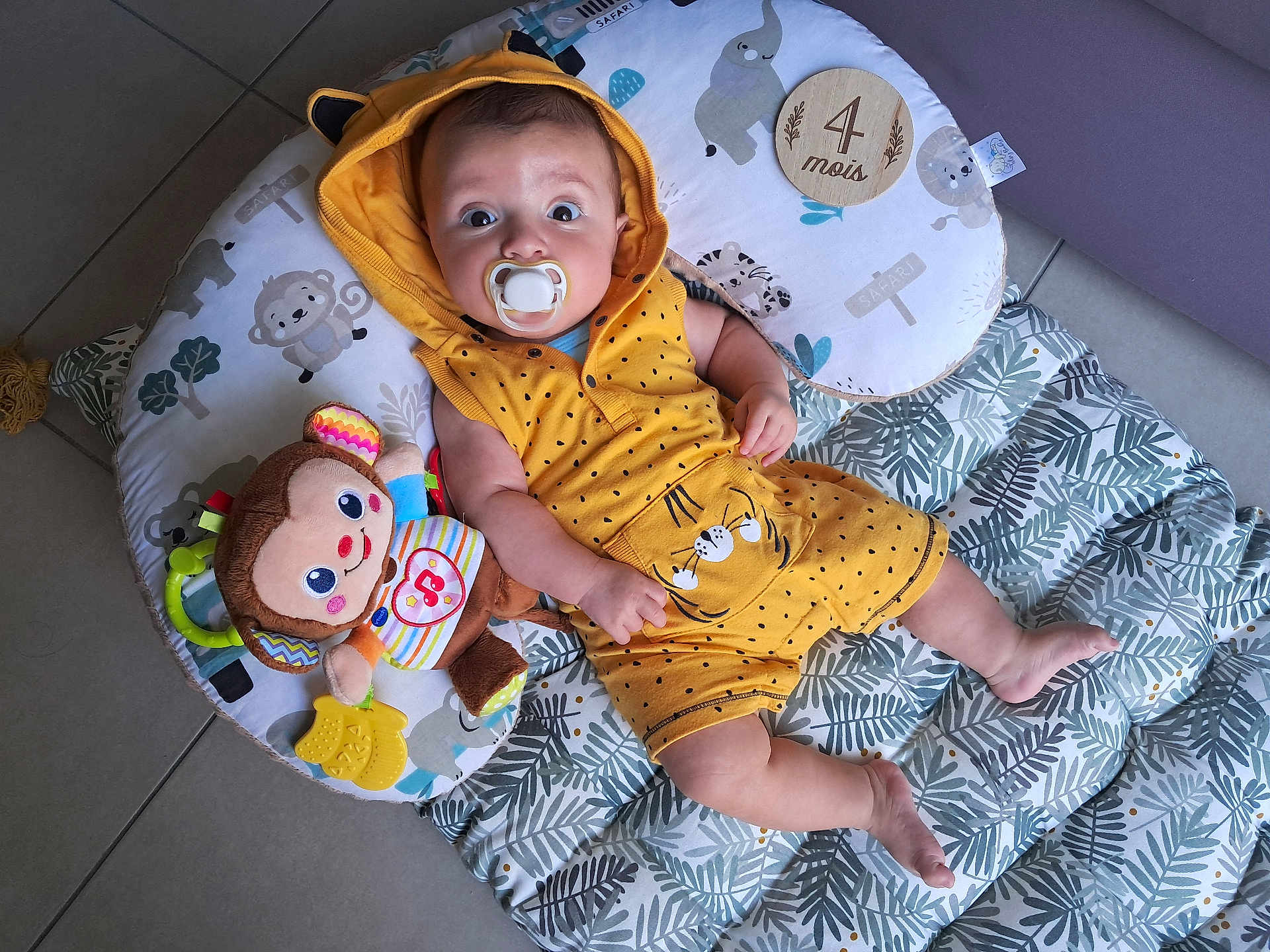 Joseph a rejoint le concours — aidez-le/la à gagner de superbes lots ! baby, infant, pacifier, yellow_clothing, hooded_romper, plush_toy, monkey_toy, floor_mat, leaf_pattern, wooden_plaque, age_marker, indoor, tile_floor, cute, child, young_child, lying_down, wide_eyes, baby_girl, baby_boy