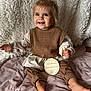 toddler, child, smiling, blue_eyes, hair_bow, brown_clothing, knit_vest, plush_blanket, soft_texture, wooden_plaque, first_birthday, sitting, indoors, happy, cute, portrait, baby, floor, cozy, celebration