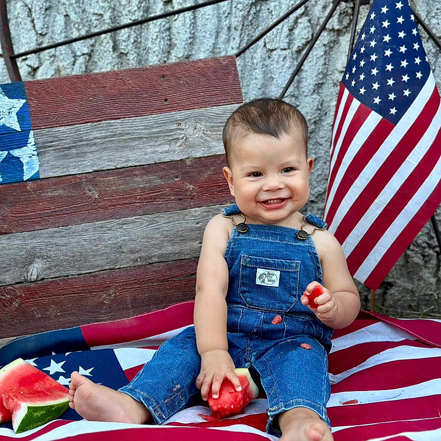 Royce is registered to the contest to win money with this photo: american_flag, bench, blue, child, coca_cola, denim_overalls, food, fruit, glass_bottle, grass, happy, outdoor, patriotic, picnic, red, smiling, summer, toddler, watermelon, white