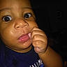 Braylin is registered to the contest to win money with this photo: baby, child, face, hand, finger, curious, dark_background, close_up, skin, eyelashes, clothing, toddler, indoors, expression, person, portrait, cute, chewing, young, wearing