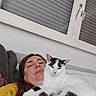person, cat, indoor, window, shutter, relaxed, resting, yellow_clothing, white_wall, brown_hair, black_and_white_cat, couch, casual, selfie, pet, comfort, close_up, home, animal, face