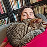 cat, tabby_cat, person, couch, bookshelf, books, indoor, relaxed, sleeping_cat, cozy, furniture, glasses, red_clothing, home, pet, nap, fur, domestic_cat, resting, comfort