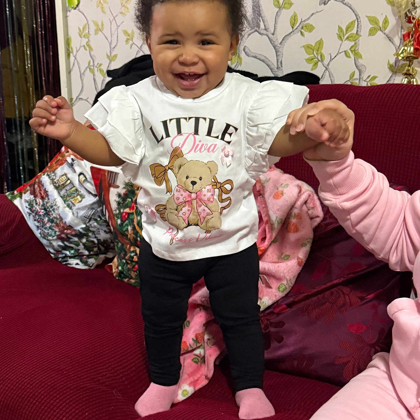 Larissa-Rose is registered to the contest to win money with this photo: black_pants, blanket, child, couch, cute, family, happy, holding_hand, home, indoor, owl_pattern, person, pink_socks, red_couch, smiling, standing, support, toddler, wallpaper, white_shirt