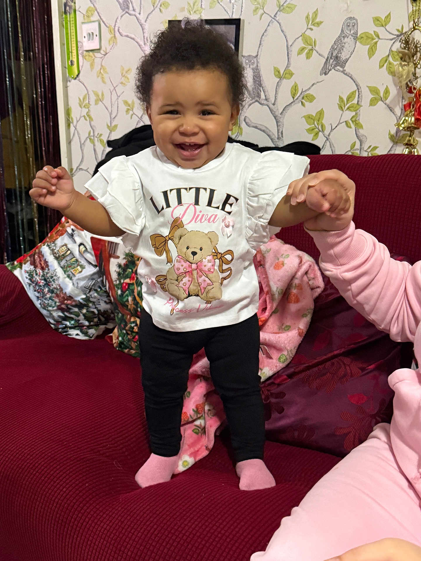 Larissa-Rose is registered to the contest to win money with this photo: toddler, child, smiling, standing, holding_hand, couch, red_couch, white_shirt, black_pants, pink_socks, blanket, wallpaper, owl_pattern, indoor, person, happy, home, cute, family, support