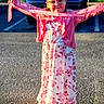 child, girl, smiling, outdoors, parking_lot, car, floral_dress, pink_clothing, cardigan, scarf, sleeves, glasses, standing, sunlight, shadow, suburban, house, fashion, portrait, playful