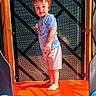 Damien joined the competition — help win amazing prizes! child, toddler, standing, smiling, portrait, outdoor, play_structure, playground, slide, colorful, sunlight, shadows, wood, mesh, blue_shirt, shorts, barefoot, face, happy, photography