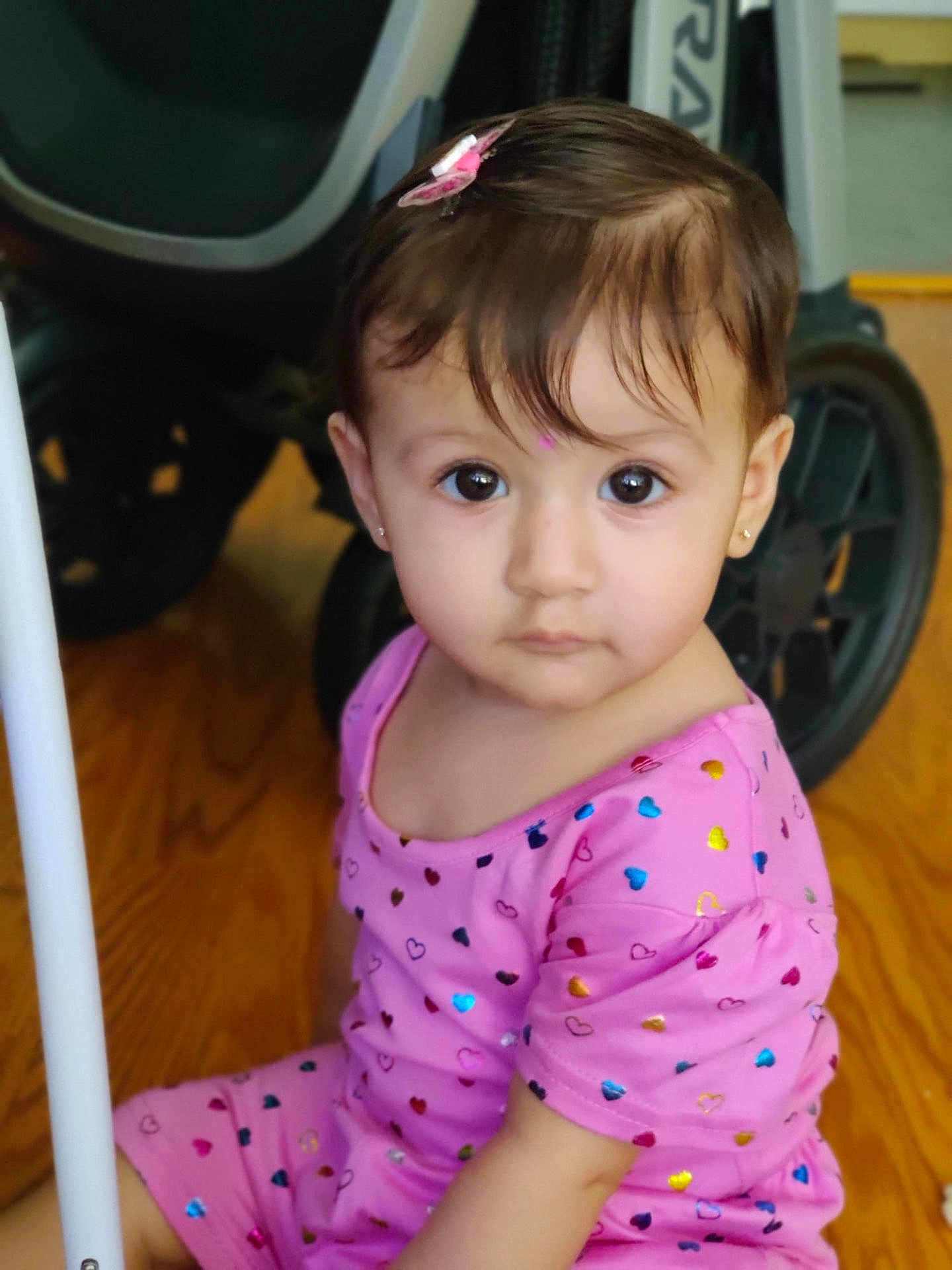 Yashvi is registered to the contest to win money with this photo: toddler, child, pink_clothing, hearts_pattern, indoor, floor, wooden_floor, stroller, wheel, face, portrait, earrings, hair_clip, curious, sitting, young_child, large_eyes, cute, person, baby
