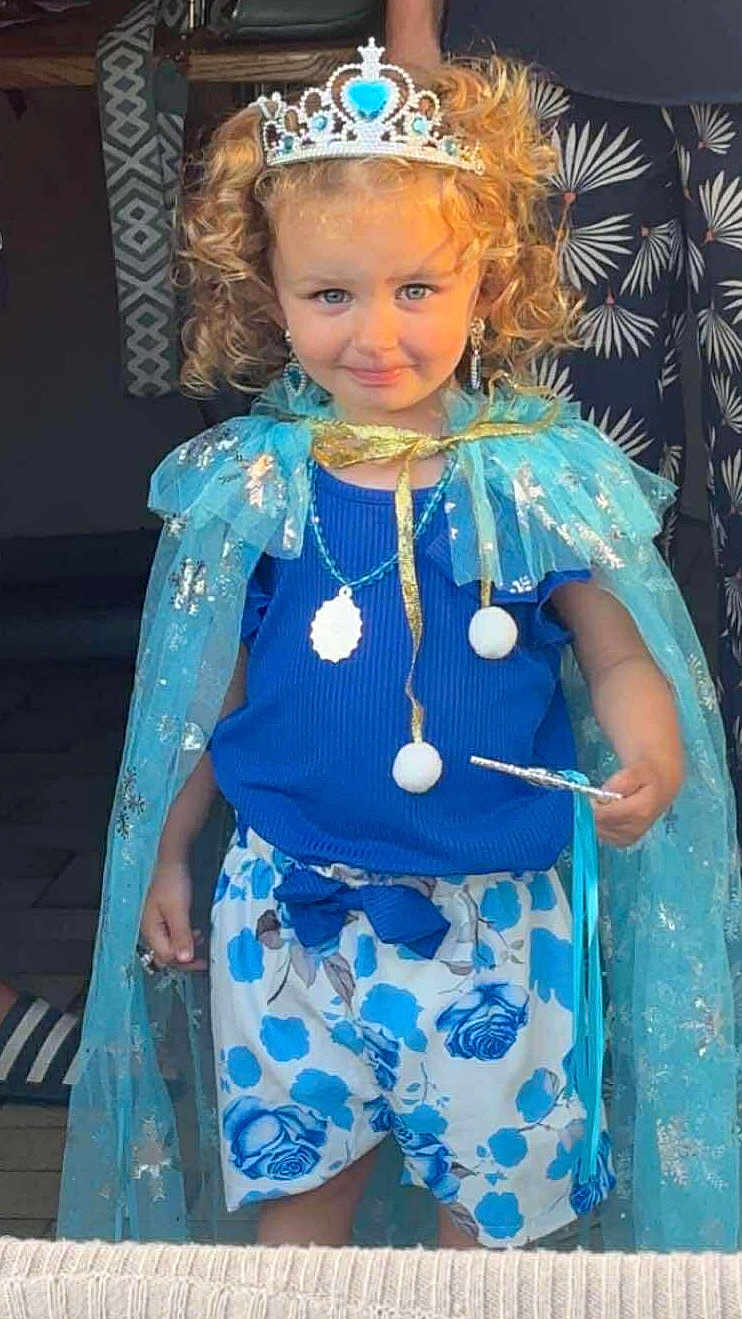 Estelle a rejoint le concours — aidez-le/la à gagner de superbes lots ! child, girl, tiara, cape, blue_clothing, shorts, flower_pattern, wand, curly_hair, smile, earrings, necklace, portrait, costume, playful, indoor, person, cute, fashion, accessories
