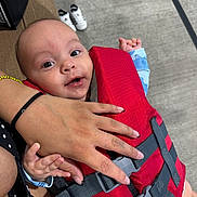 Jayden joined the competition — help win amazing prizes! baby, child, life_jacket, hand, polished_nails, drool, curious, cheerful, couch, carpet, baby_shoes, red, blue_clothing, person, indoor, close_up, holding, sitting, infant, smiling