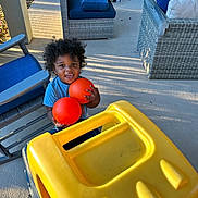 Carson joined the competition — help win amazing prizes! toddler, child, toy, orange_balls, yellow_toy_truck, outdoor, patio, curly_hair, blue_cushions, wicker_chair, sunlight, smiling, playing, concrete_floor, furniture, happy, daylight, person, casual_clothing, shadow