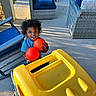 toddler, child, toy, orange_balls, yellow_toy_truck, outdoor, patio, curly_hair, blue_cushions, wicker_chair, sunlight, smiling, playing, concrete_floor, furniture, happy, daylight, person, casual_clothing, shadow