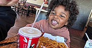 Carson is registered to the contest to win money with this photo: child, toddler, curly_hair, smiling, food, noodles, drink, cup, restaurant, table, adult, happy, indoor, casual_dining, wall_art, wooden_chairs, brown_hoodie, meal, candid, people