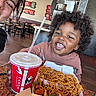 child, toddler, curly_hair, smiling, food, noodles, drink, cup, restaurant, table, adult, happy, indoor, casual_dining, wall_art, wooden_chairs, brown_hoodie, meal, candid, people