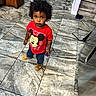 Carson is registered to the contest to win money with this photo: toddler, child, indoor, floor_tiles, red_shirt, mickey_mouse, curly_hair, slippers, toy, looking_up, wooden_cabinet, pet_bowls, pants, person, home, casual, expression, standing, young_child, curious