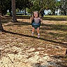 child, toddler, girl, swing, playground, outdoor, park, smiling, portrait, face, striped_shirt, shorts, shoes, socks, trees, pine_trees, sand, grass, houses, sunlight