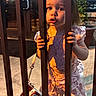 child, toddler, girl, portrait, face, blue_eyes, blonde_hair, dress, frilly_clothing, standing, hands, bars, fence, outdoor, evening, warm_lighting, curious, serious_expression, playground, photography