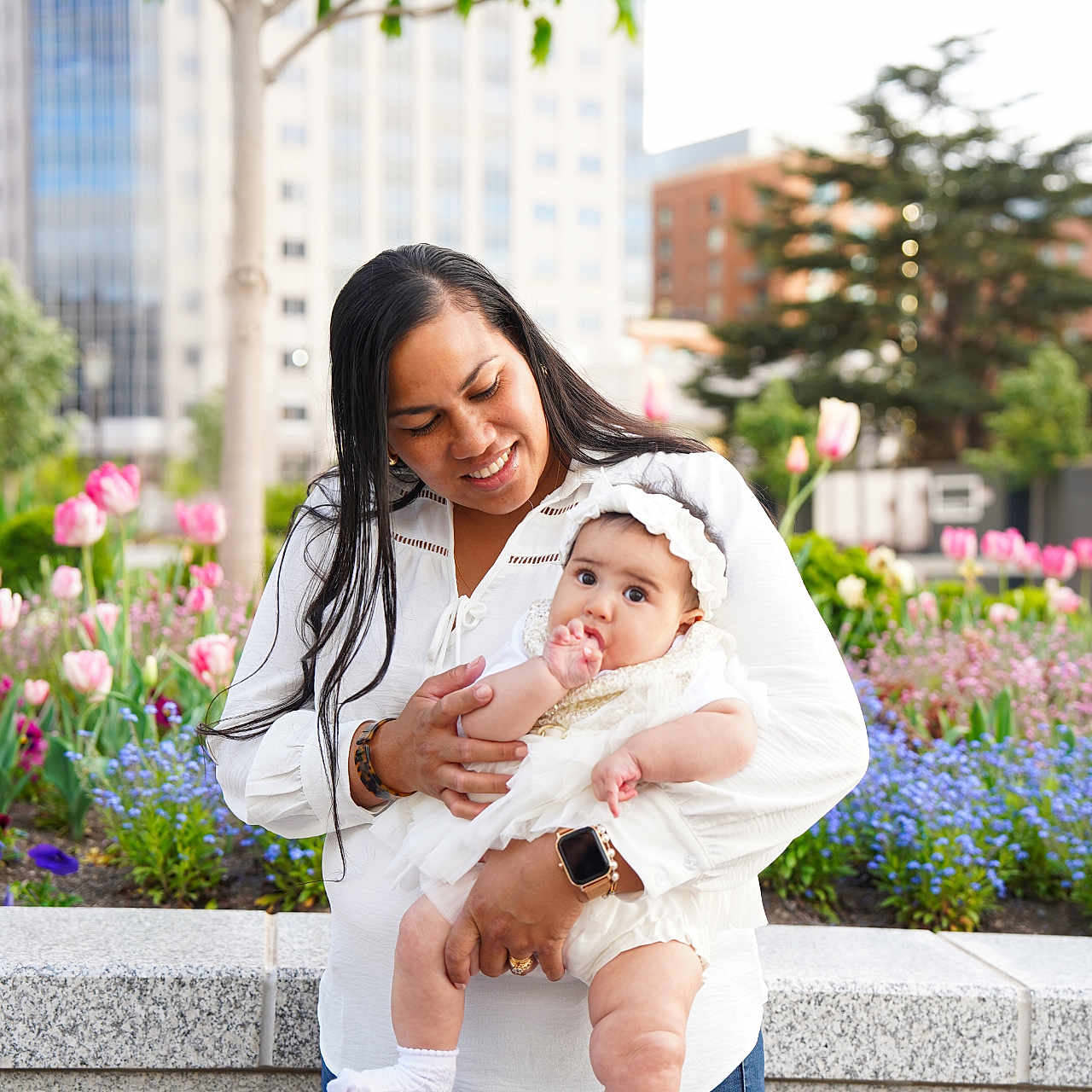 Alice is registered to the contest to win money with this photo: baby, mother, child, infant, portrait, outdoor, garden, flowers, tulips, city, urban, family, parenting, smiling, white_dress, white_clothing, headband, socks, trees, buildings