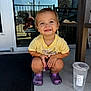toddler, child, smiling, yellow_shirt, purple_crocs, squatting, concrete, porch, plastic_cup, water, glass_door, reflection, person, outdoor, sunlight, short_hair, casual_clothing, happy, cute, daytime