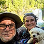 person, dog, waterfall, forest, nature, smiling, outdoor, hiking, glasses, hat, backpack, happy, curly_hair, nose_ring, tongue_out, white_dog, sunlight, greenery, portrait, travel