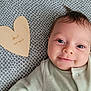 baby, infant, smiling, face, eyes, hair, blanket, pattern, wooden_heart, text, clothing, cute, portrait, lying_down, indoors, baby_garment, skin, closeup, happy, two_months