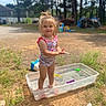 child, toddler, swimsuit, water, plastic_tub, outdoor, sunny, grass, sand, toys, basketball_hoop, trees, smiling, standing, sandals, play, summer, young, happy, person