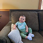 Leland is registered to the contest to win money with this photo: baby, smiling, couch, pillow, indoor, child, infant, clothing, socks, happy, face, person, furniture, home, wood, window, cozy, seated, cute, relaxed