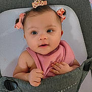 Eliana is registered to the contest to win money with this photo: baby, child, infant, pink_bib, hair_accessories, crown_clip, baby_carrier, grey_fabric, cute, adorable, smiling, small_hands, face, portrait, indoor, closeup, happy, young_child, person, seat