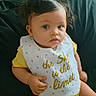 Eliana joined the competition — help win amazing prizes! baby, child, curly_hair, yellow_clothing, bib, text_on_bib, sitting, indoor, person, cute, face, expression, hand, skin, young_child, infant, closeup, portrait, soft_lighting, comfortable