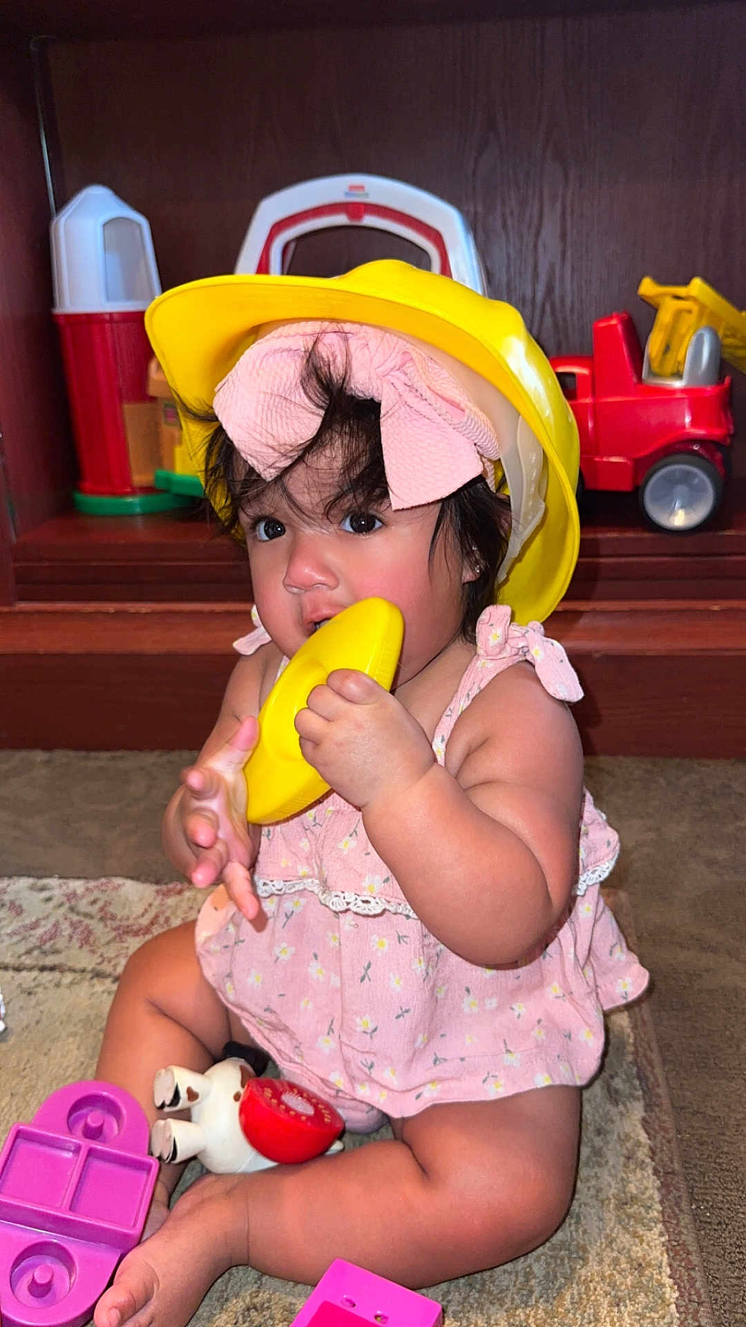 Liliana is registered to the contest to win money with this photo: toddler, child, yellow_helmet, pink_dress, bow, toy, plastic_toy, carpet, indoor, play, cute, baby, sitting, curious, wooden_shelf, background, colorful, hand, face, person