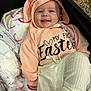baby, child, hoodie, smiling, tongue_out, blanket, diaper, clothing, cute, infant, happy, person, indoors, laying_down, soft_toys, pastel_colors, celebration, easter, white_pants, beige