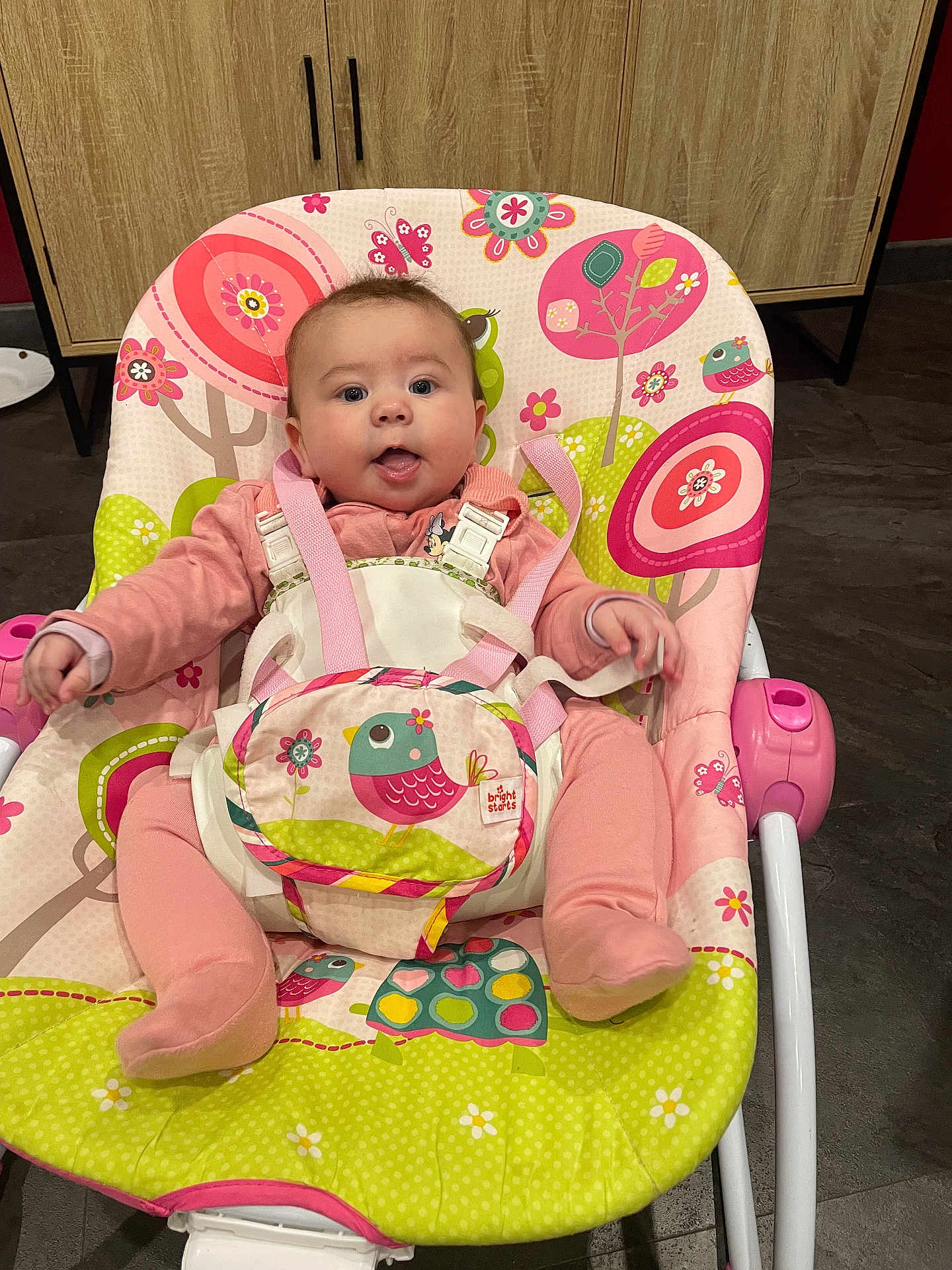 Yüna a rejoint le concours — aidez-le/la à gagner de superbes lots ! baby, child, pink_outfit, baby_rocker, colorful, bird_pattern, flower_pattern, indoor, cute, face, tongue_out, seat, straps, floor, cabinet, wooden_cabinet, playful, infant, happy, person