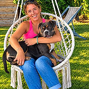 Nala participe au concours pour gagner de l'argent avec cette photo : woman, dog, pet, outdoor, garden, lawn, hanging_chair, smiling, portrait, summer, sunlight, greenery, backyard, sunglasses, pink_shirt, blue_jeans, tattoo, bracelet, relaxed, companion