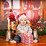 Noé participe au concours pour gagner de l'argent avec cette photo : child, baby, toddler, santa_hat, gift_box, present, christmas, holiday, elf_doll, wooden_floor, bells, pinecone, wreath, festive_decor, smiling, portrait, indoor, sitting, toy, decorations