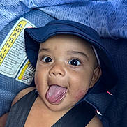 Liam joined the competition — help win amazing prizes! baby, child, face, hat, tongue, car_seat, seatbelt, warning_label, closeup, expression, playful, cute, infant, headwear, strap, person, skin, indoors, young, smile
