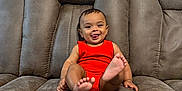 Liam is registered to the contest to win money with this photo: baby, child, smiling, tongue_out, red_clothing, sitting, couch, brown_couch, indoor, happy, cute, plush_furniture, person, infant, comfortable, playful, home, casual, portrait, furniture