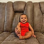 Liam is registered to the contest to win money with this photo: baby, red_onesie, couch, brown_couch, smiling, tongue_out, infant, sitting, indoor, plush, cute, child, happy, person, furniture, skin, foot, hand, head, playful